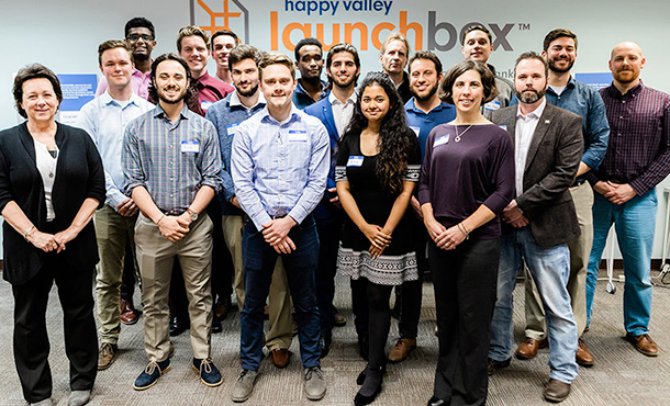 18 students and professors stand and pose for a group picture in front of a wall that says "Happy Valley LaunchBox"